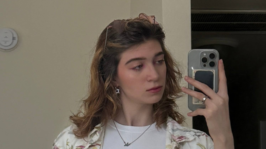A young girl takes a selfie in the mirror on her smartphone. She is wearing a white T-shirt and a shirt with a floral pattern, her hair is slightly wavy, and she is wearing sunglasses. She is looking away, her expression is calm.